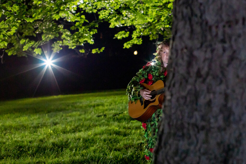 A night time photograph of a grassy landscape viewed from under the canopy of a large tree. A musician dressed in a leafy costume plays a guitar from behind the tree trunk in the foreground at right.