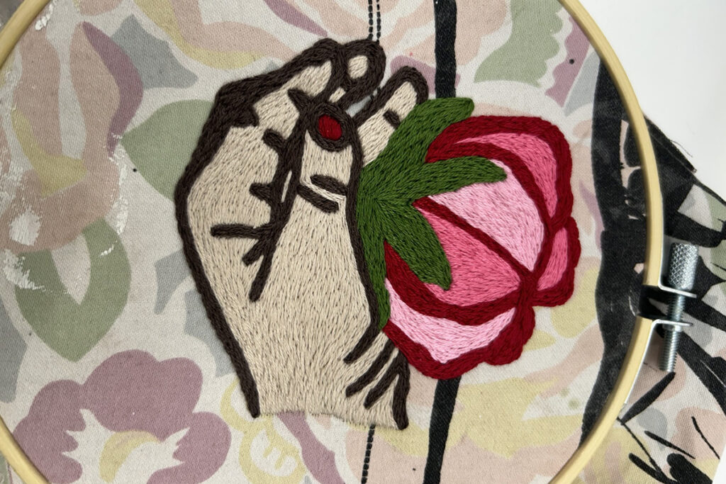 A swatch of fabric with a floral pattern features an embroidered drawing of a hand holding a rosebud. Overtop the fabric is a wooden hoop encircling the embroidery.