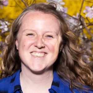 A portrait of the composer and musician Emily Bate, a smiling white woman with dark-blonde wavy hair. She is outdoors with autumn leaves appearing in a tree nearby.