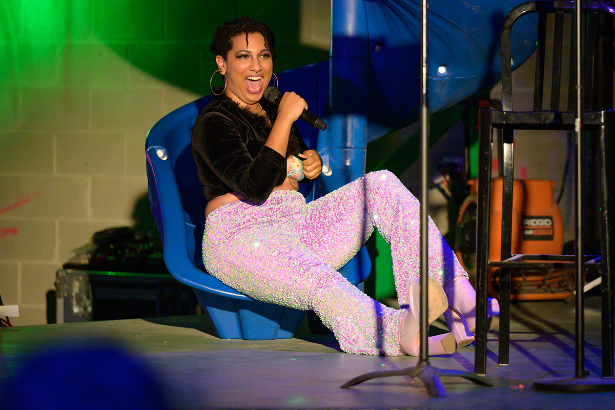 A photo of a person, Jackie Soro, singing into a mic while semi-reclined in a low chair on a stage. They are wearing a black top and sparkly pink pants.