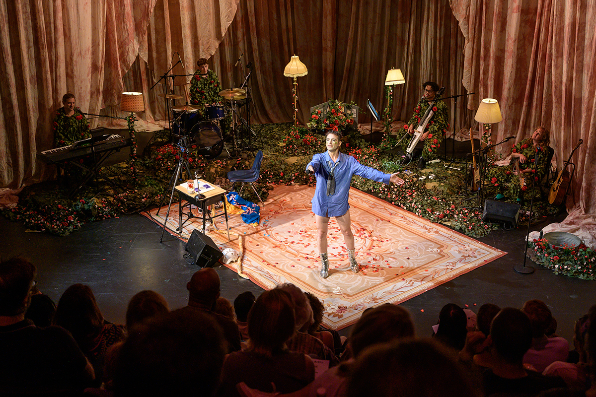 A photo of a band performing on stage in front of a live audience. A singer stands on a carpet wearing an oversized blue dress shirt and tie, extending the mic toward the audience. In the background, surrounding the rug, are four musicians wearing outfits composed of vines and roses. They sit with their instruments between four upright lamps.