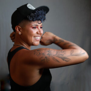 A portrait of Be Steadwell, a queer person of color with short curly dark hair. They are smiling at a three-quarter pose with their arm flexed. They are wearing a baseball cap and black tank top. 