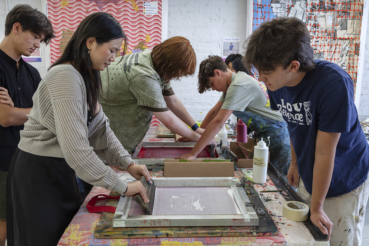 Six teens gather around a table and work together to print art using silkscreens.