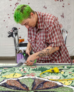 An artist, Bailey Roper, works with their hands on the back-side of a rug. They have short green hair and are wearing a red flannel shirt.