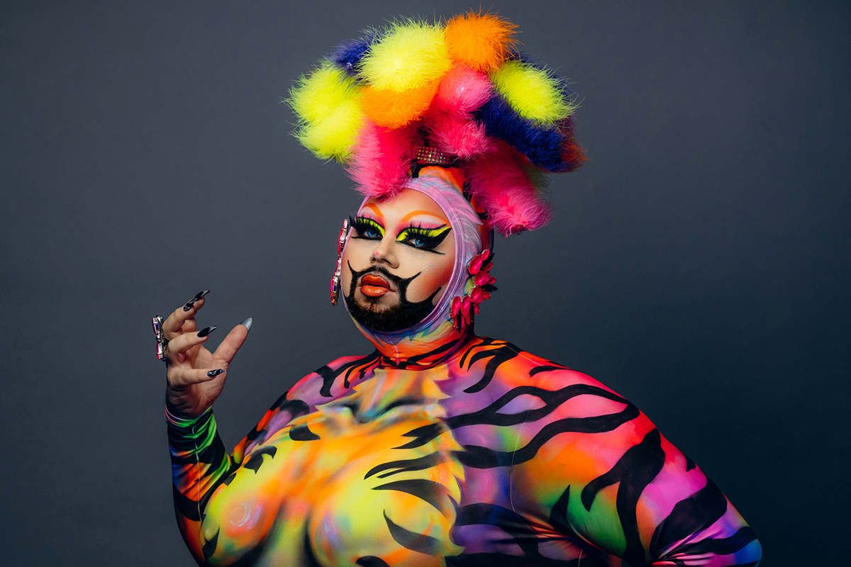 A drag artist, Eric Jaffe, wears a technicolor outfit with tiger stripes and a headpiece consisting of tufts of bright colored fur. Her beard is highly stylized, accenting her make-up composed of saturated colors.