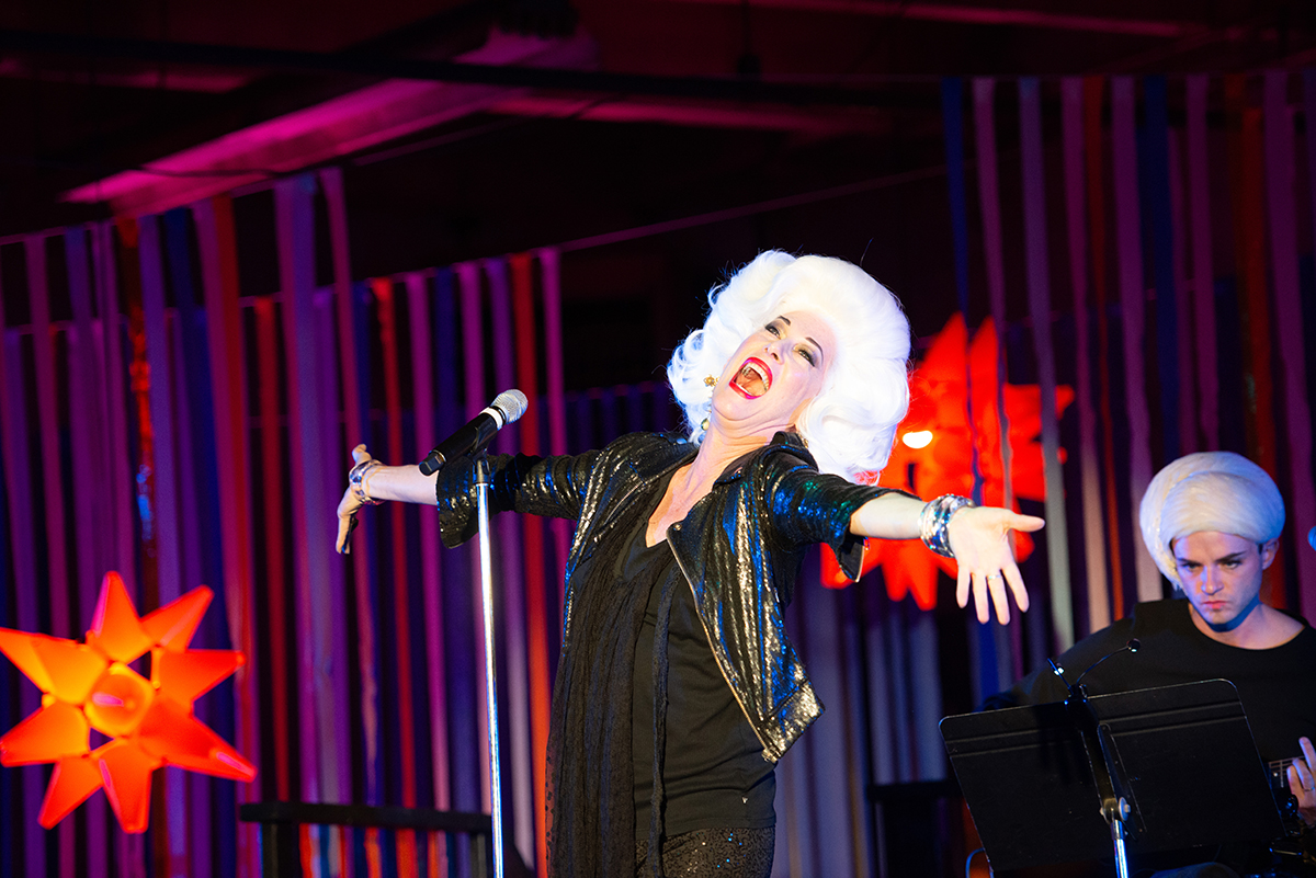 A performer, Justin Vivian Bond, sings with arms outstretched on a stage. They are wearing a black leather jacket and a large white wig.