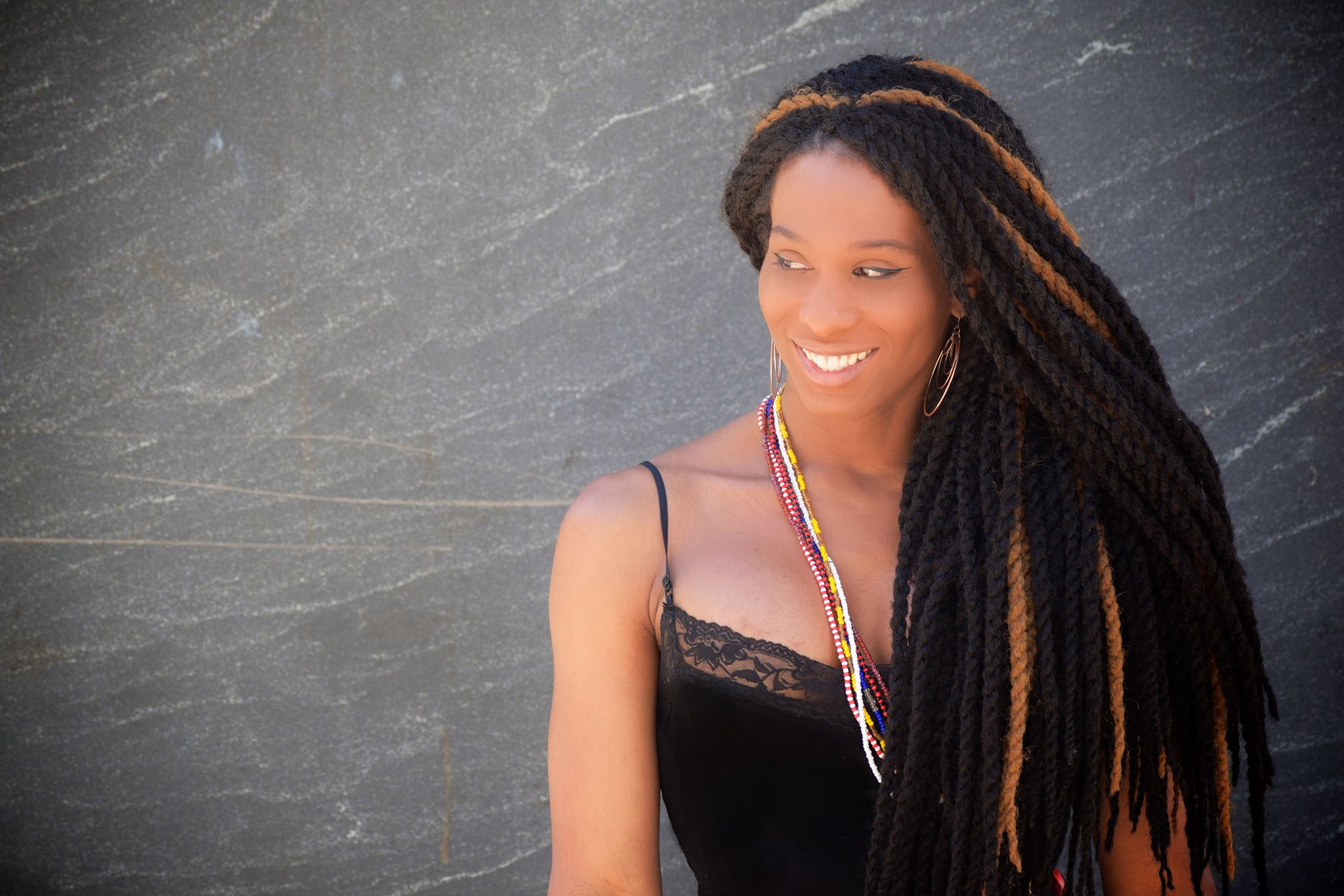 A portrait of Lady Dane, a Black trans woman who is seated against a gray wall. She has long braided hair and is wearing a black dress with shoulder straps and a necklace. She is smiling and looking to her right.