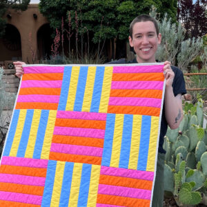 A picture of a white person smiling while holding a quilt in front of them. The quilt consists of a grid of nine squares with the corners and center square pink and the others a series of vertical blue and yellow stripes.