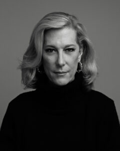 A black and white portrait of Justin Vivian Bond, a white trans woman with stylish blonde hair that sits just above her shoulders. She is wearing a black turtleneck and hoop earrings.