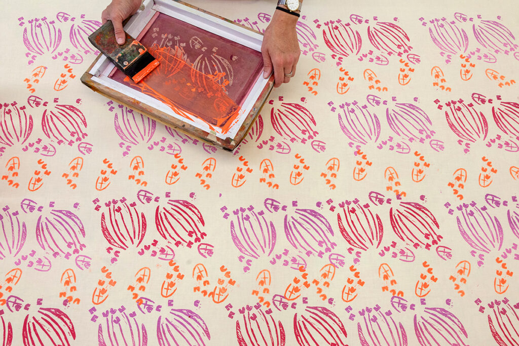 A picture of a repeating pattern of alternating floral shapes printed in warm colors. At the top of the picture, someone's hands appear holding a silkscreen and a small squeegee dragging orange ink across its surface.