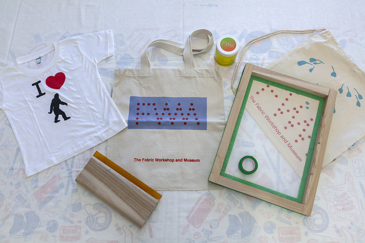 A photo of an array of materials that comprise a screenprinting kit: a graphic t-shirt, a squeegee, totes, a silkscreen, and a jar of ink.