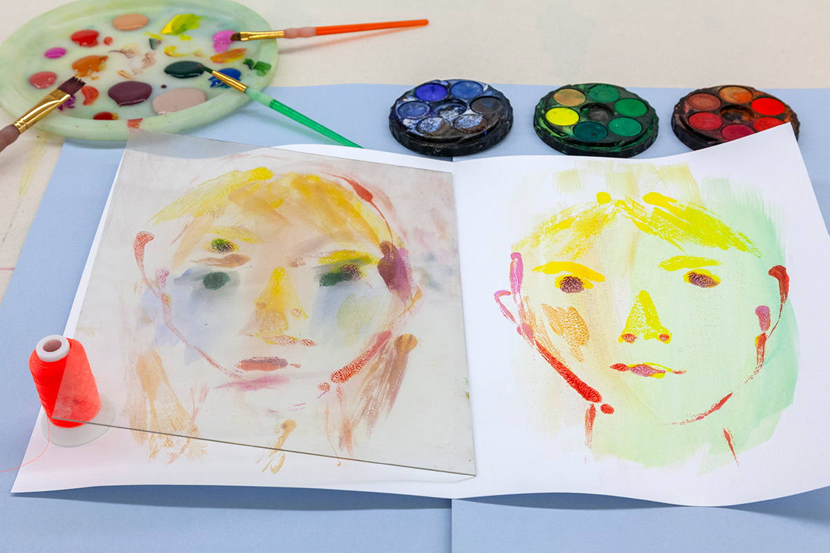 A folded sheet of paper with a watercolor portrait on each page sits on a table near brushes and a watercolor palette.