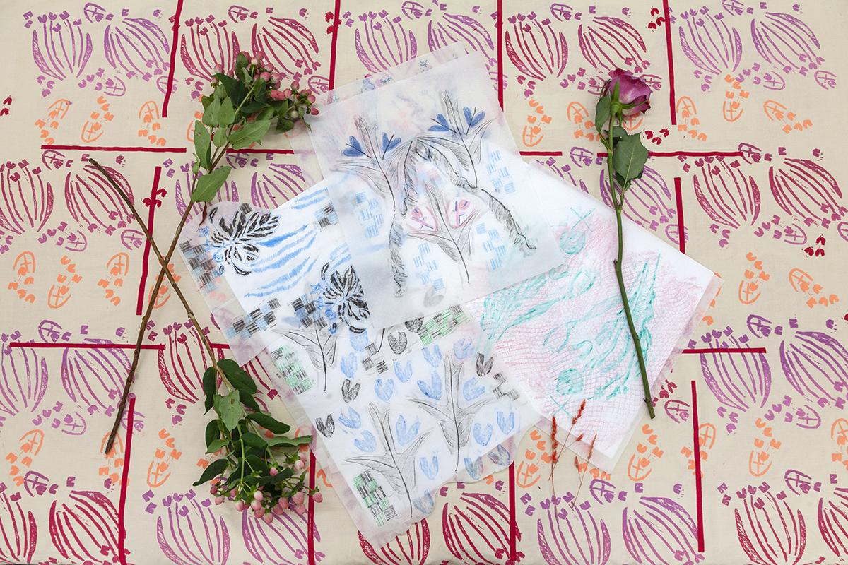 Sheets of floral-inspired drawings lie upon a surface featuring a repeating pattern of other floral motifs printed in warm colors. Rose cuttings accent the edges of the paper drawings.