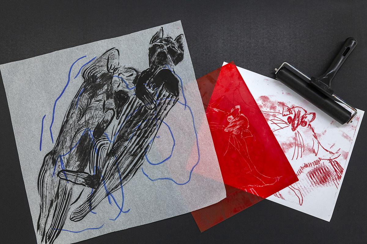 A black drawing of two cats stretching on a semi-transparent sheet of paper lies atop sheets of red and white paper with a print of another cat drawing. A small print roller rests near the white paper.