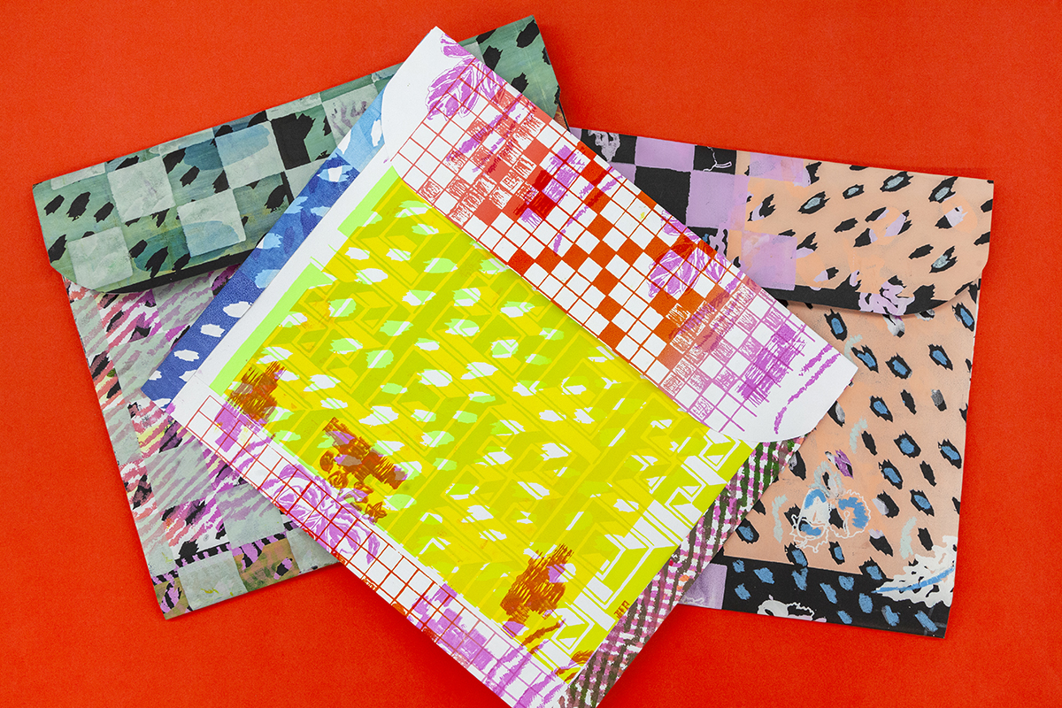 Three trapper-keeper style folders lie against a red surface. The folders feature wild colorful patterns of checkerboard grids and drawings.