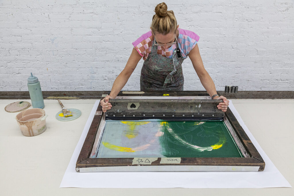 A photo of a white woman screenprinting using a squeegee to push ink through a silkscreen.