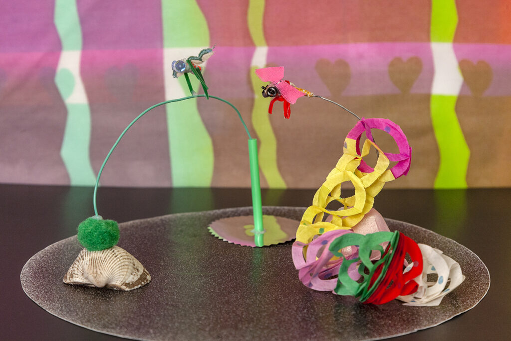 A photo of a miniature set of sculptures resembling a circus trapeze. Small sculptural fleas made of fabric animate the set.