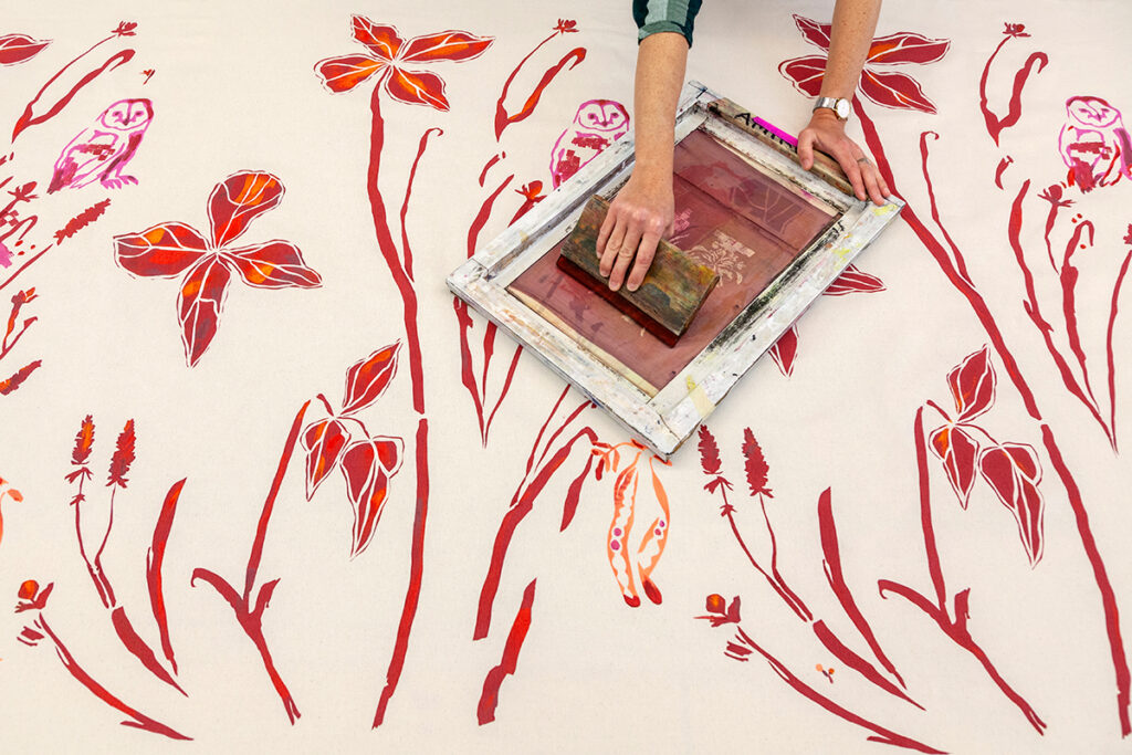 A hand reaches across a table to drag a small squeegee across a silkscreen to print a floral design in a warm, fall palette on cream linen fabric.