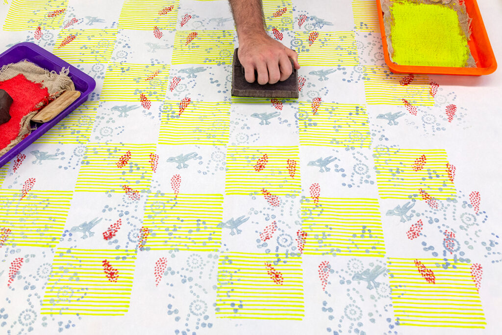 A hand reaches across a printed surface to stamp a pattern using a woodblock. The resulting pattern is a yellow and white checkerboard pattern with overlaying textures.