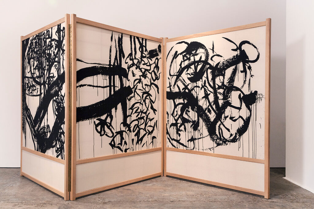 A large, tri-fold room divider made of light wood and cream fabric. On the fabric there are large, abstract gestural marks in black.