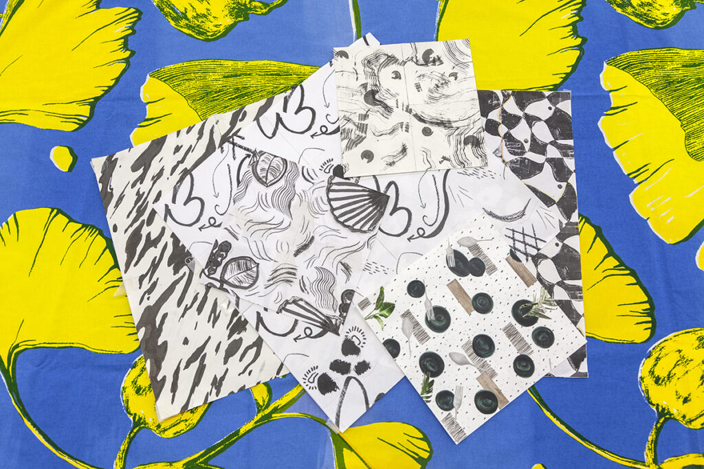 An array of black and white drawings with varying patterns lie across a blue surface with patterns of yellow ginkgo leaves.