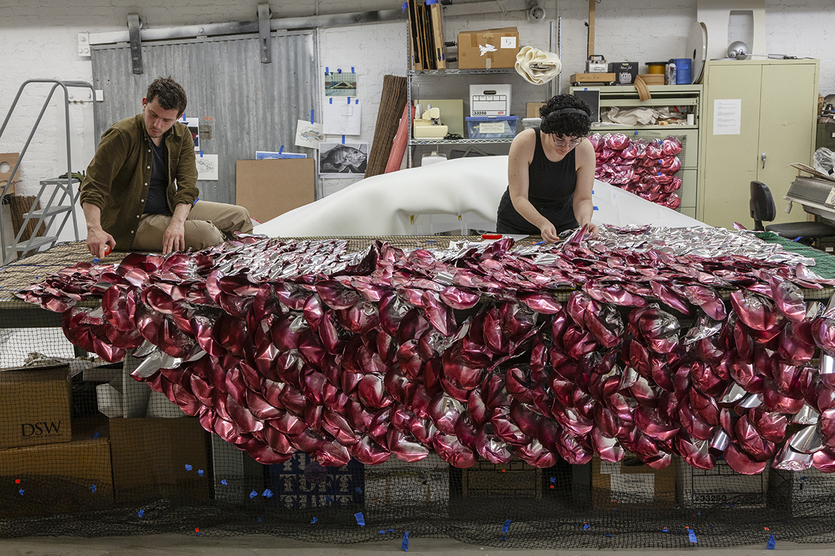 Two artists work in a studio to fit red-painted scales into a mesh fabric.