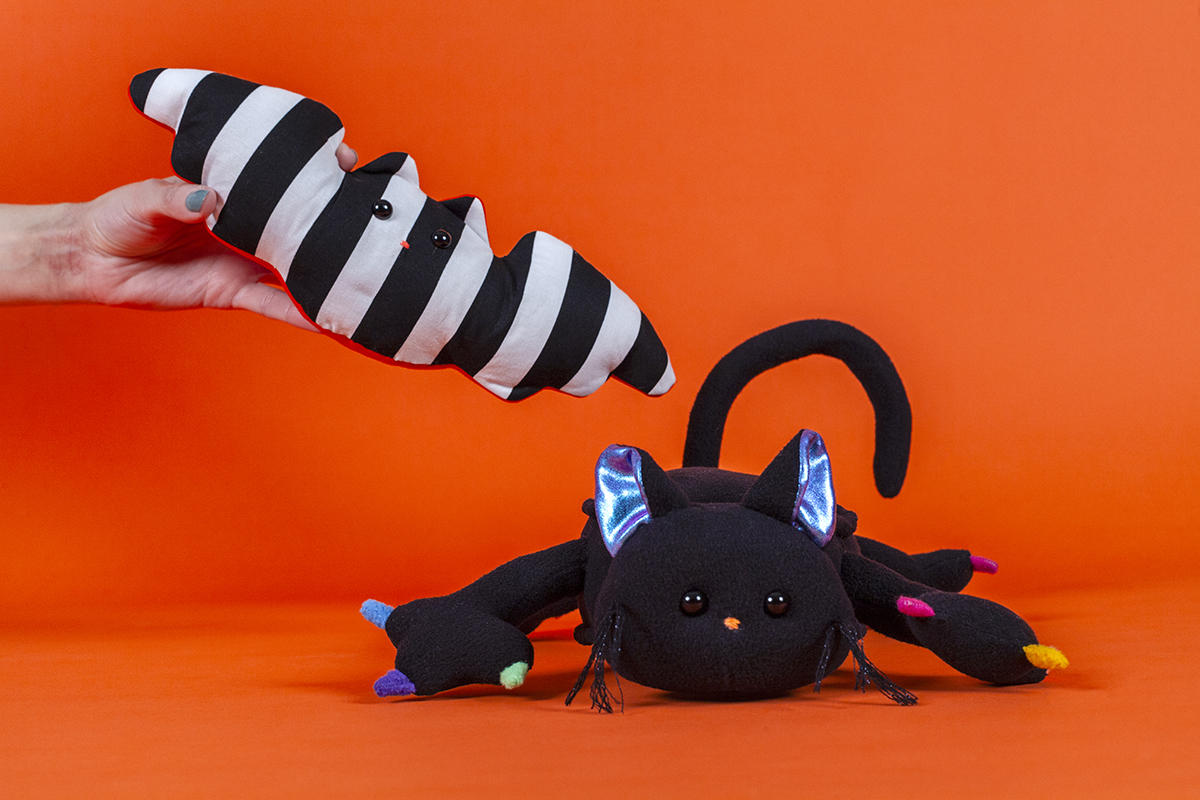 A picture of two spooky plush animals—a striped bat and a black cat—against an orange background.