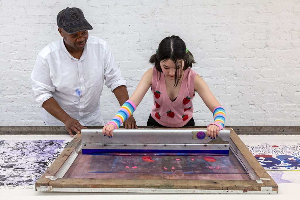 A teenage girl uses a squeegee to print on a silkscreen with guidance from an instructor.