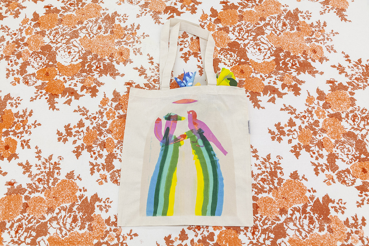 A cream-colored tote features a colorful print of a bird and cactus flower. The tote lies across a broader surface with a repeat floral pattern printed with burnt orange ink.