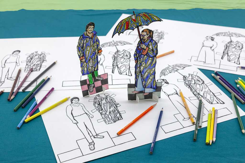 Two paper dolls stand upright on the sheets of paper that bear that outlined template. Colored pencils used to color in the dolls are scattered nearby.