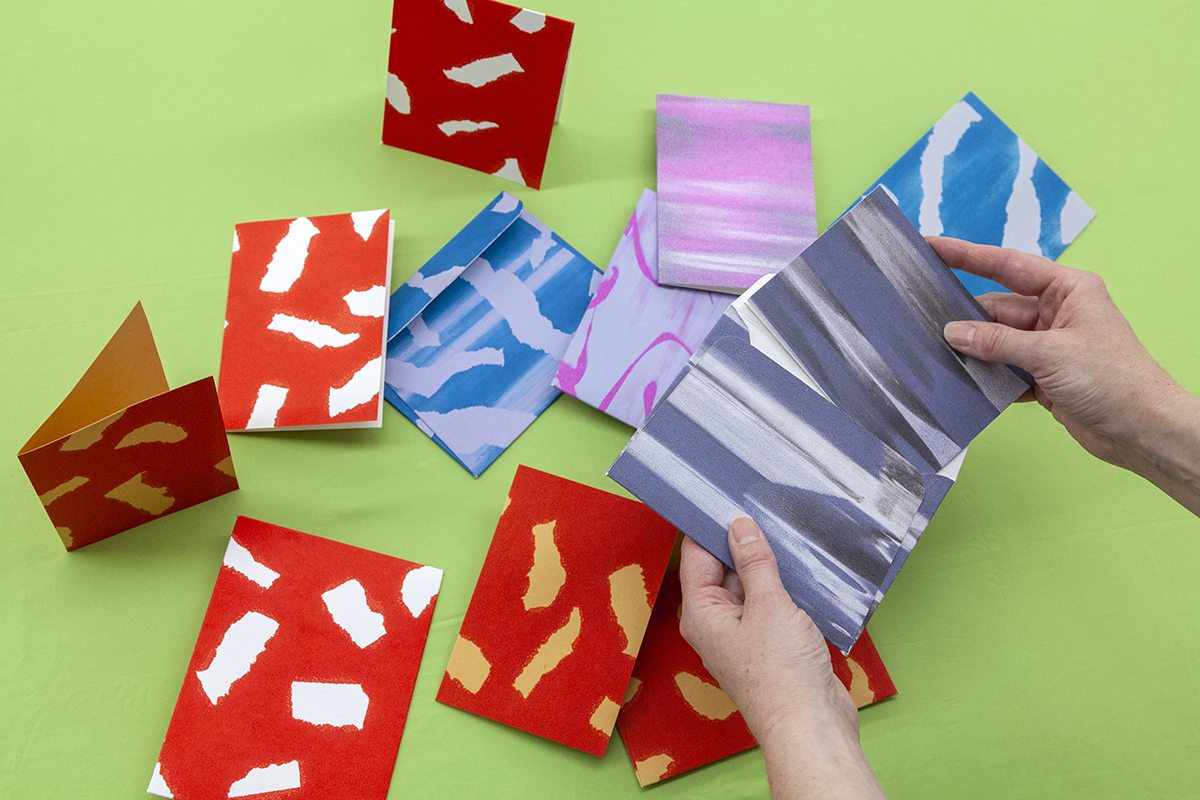 A few sets of cards are scattered across a surface, consisting of a few different abstract patterns in red, blue, and pink. A pair of hands hold one open on the right.