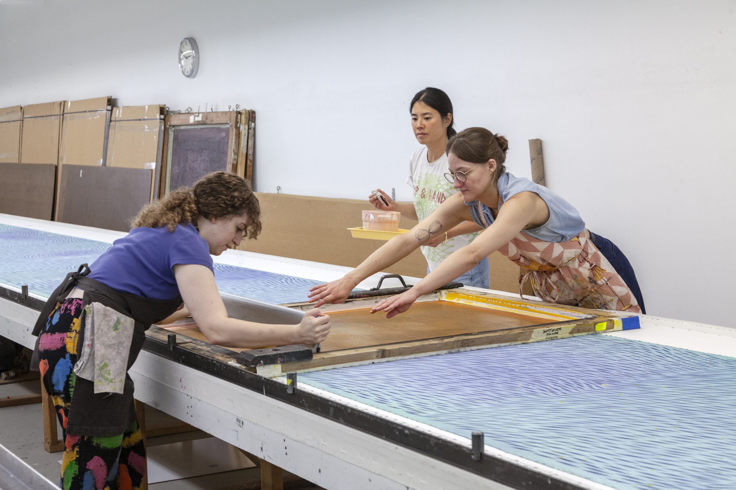 Artist Julia Chiang overseeing two screenprinters printing a repeat pattern on with a large-scale screen on a long table.