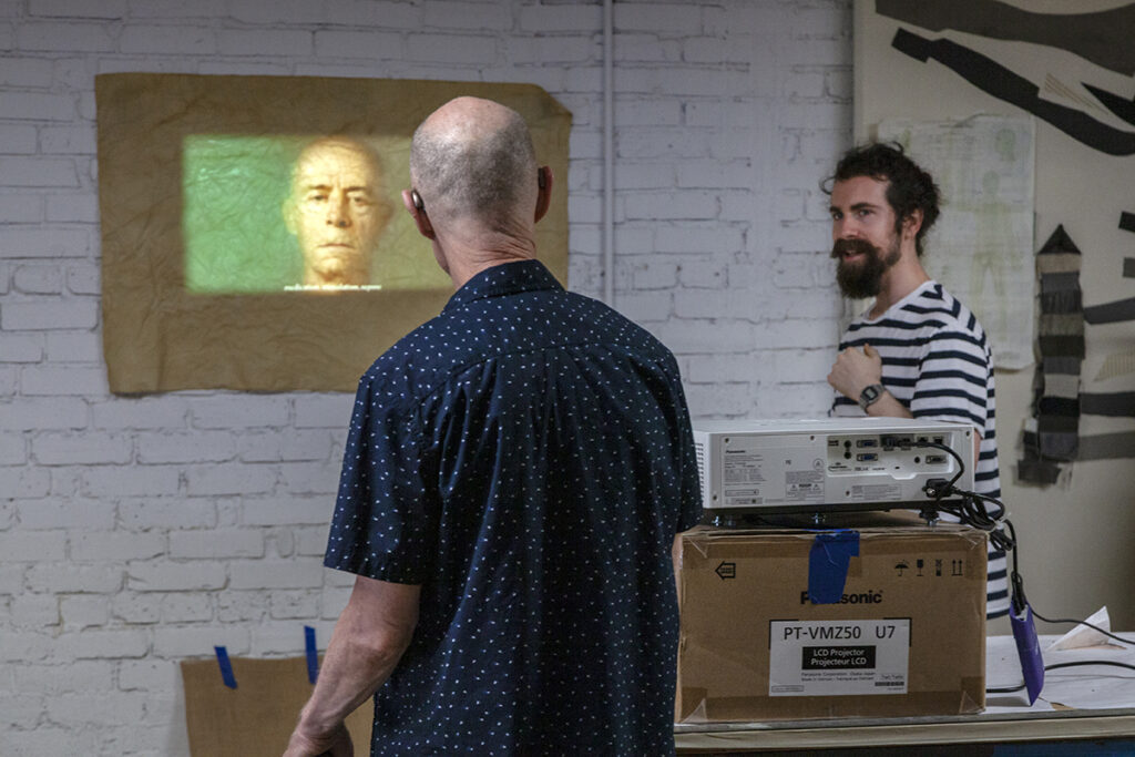 A photo of the artist John Killacky, an older balding white man, turned away from the camera and facing a projection of his own face on an artwork on the wall. At his right, a studio artist with dark hair and a beard looks on.