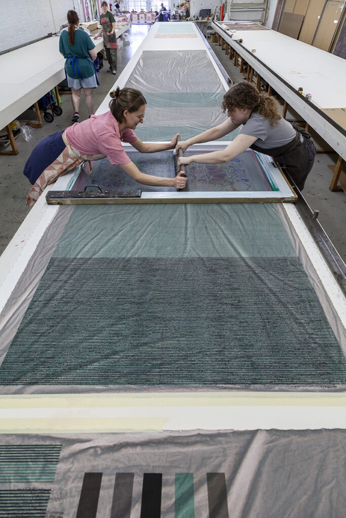 Two woman artists screenprint from either side of a large table. They are printing a fabric pattern made of dense and textured lines of gray and green.