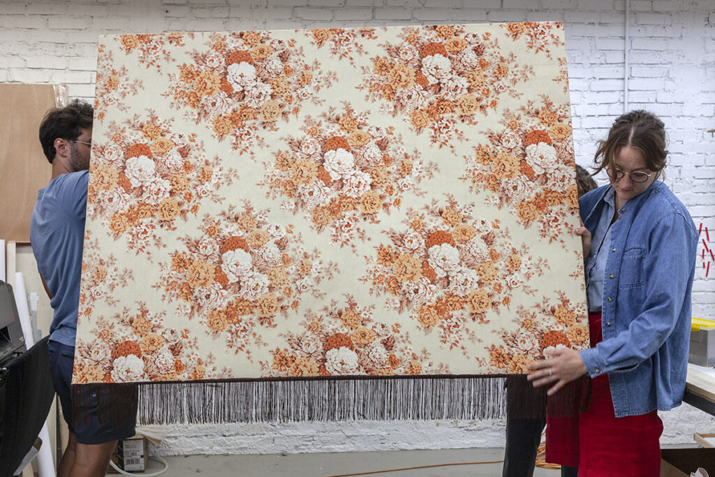 Two people carry a large suface featuring a repeated pattern of diamond-shaped groupings of florals, printed in orange and cream hues.