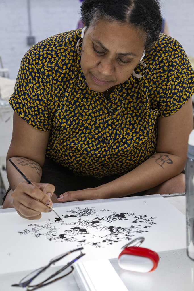 A Black woman artist leans over a table to paint flowers with black ink.