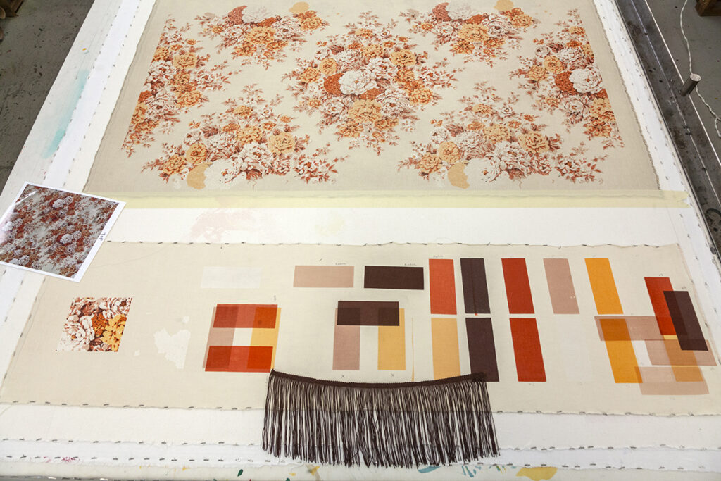 A sampling of imagery laid out on a table, including a repeated pattern of florals, printed in orange and cream hues, color swatches of each of those colors, and a sample of fringe.