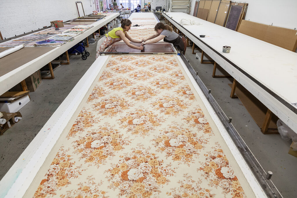 Two artists screenprint from either side of along table. They are printing a repeated diamond-shaped grouping of flowers in orange and cream hues.