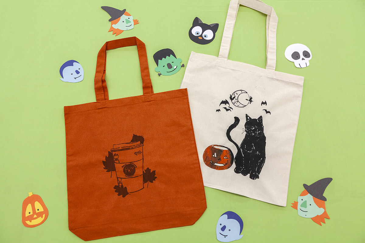 On a light green background, there are two totes laying flat. The tote on the left is burnt orange with a brown screenprint of a coffee cup surrounded by leaves. The tote on the right is cream with a screenprinted orange pumpkin and black cat.