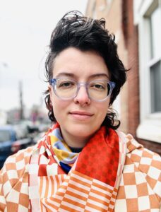 A portrait of Ash Garner, a young white person wearing a scarf with a checkerboard pattern. She wears glasses, has quaffed dark hair, and is standing outdoors.