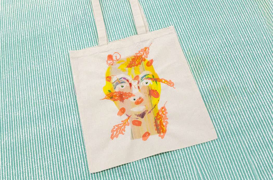 A cream-colored tote lies against a mint green background. The tote features a printed portrait of a blonde person with orange leaves overlaid, obscuring the person's face.