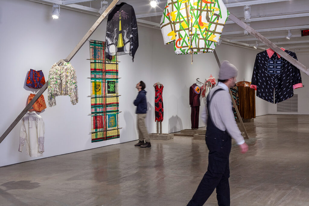 A gallery installation photo showing five garments installed on a triangular armature with other garments and artworks installed on the wall nearby.