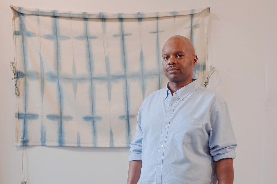 A portrait of the artist Anthony Folks, a young Black man with a shaved head. He is wearing a light blue dress shirt with rolled sleeves. He is standing in front of an indigo sample.