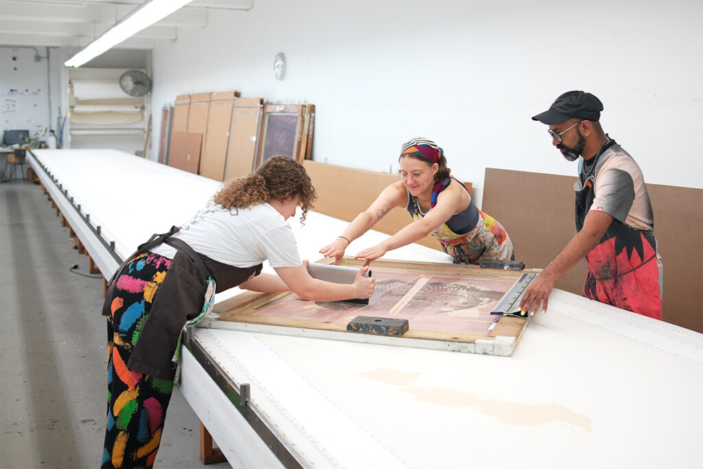 Two women artists screenprint by pushing a squeegee across a silkscreen from either side of a very long white table. A man in a black cap supports them by holding the edge of the screen in place.