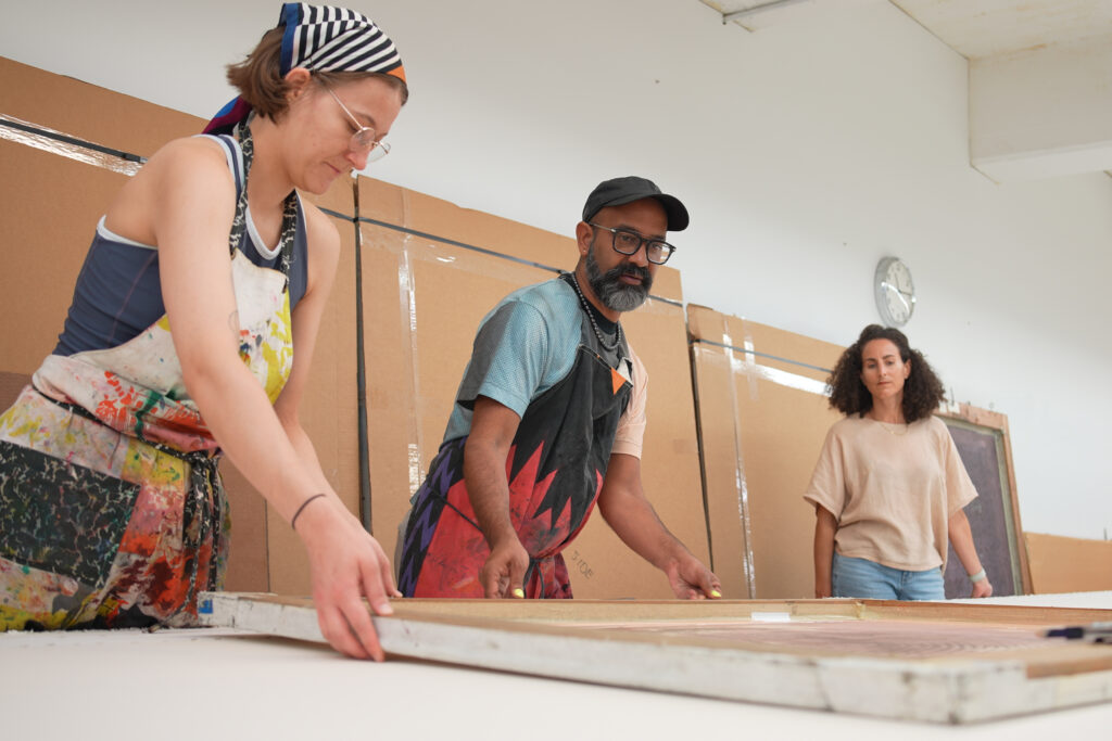 A photo taken at a low vantage point shows three people on the other side of a table. The man in the center moves a silkscreen with the woman on the left. The woman on the right observes.