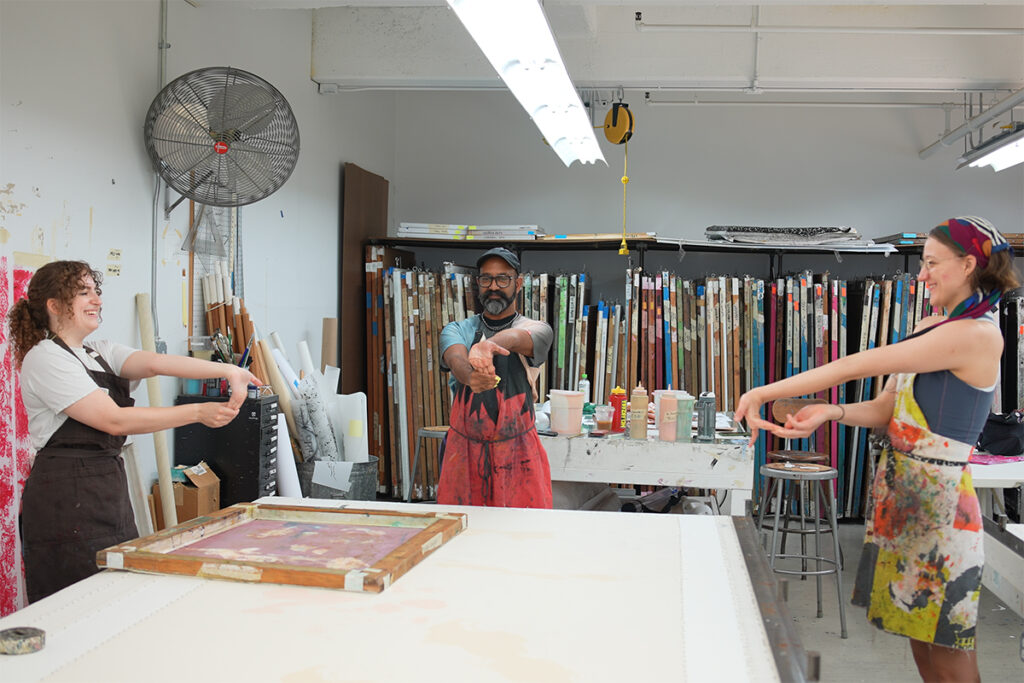 Three artists stretch their arms forward in front of their bodies, clasping their fingers together. They are positioned around three ends of a long white table in a printmaking studio..