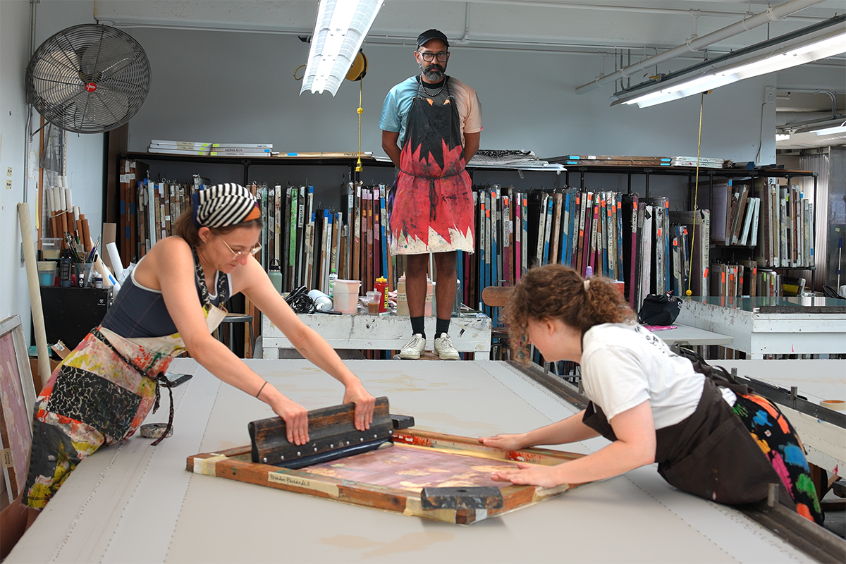 A photo of two women artists screenprinting over a long table while another artist, Brendan Fernandes, stands atop a stool in order to observe from a high vantage point.