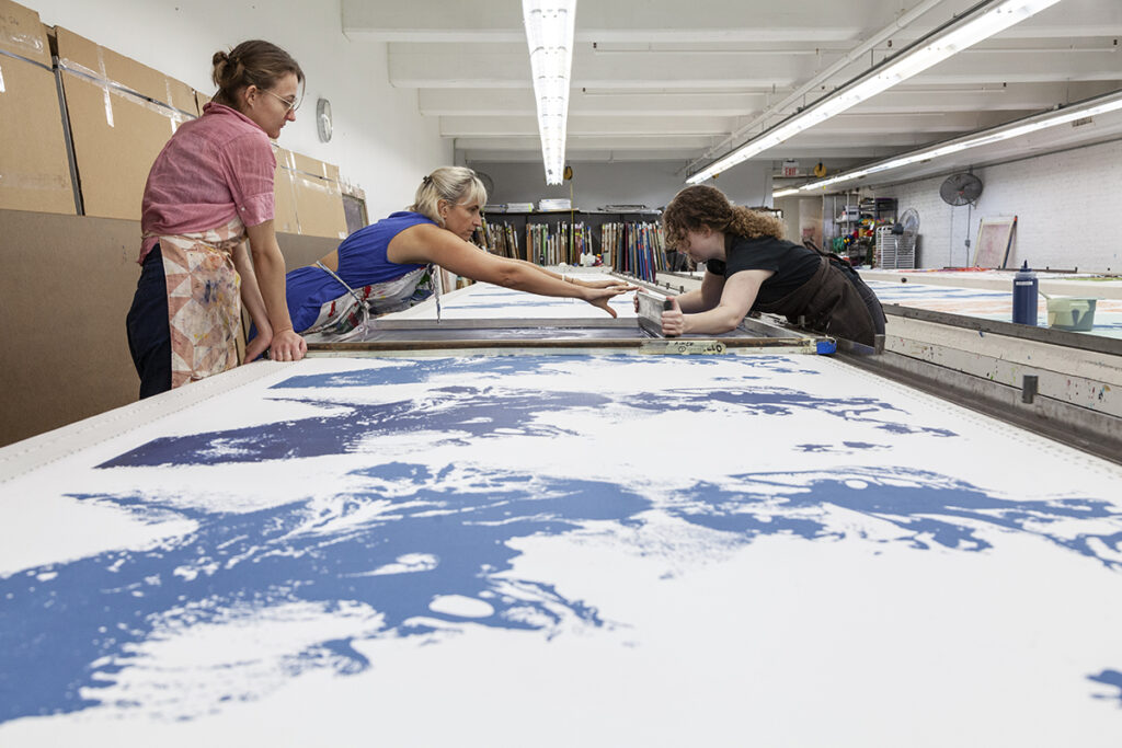 A photo of two women artists screenprinting from either side of a large table. They are printing an abstract splash-like pattern in blue while a coworker looks on.