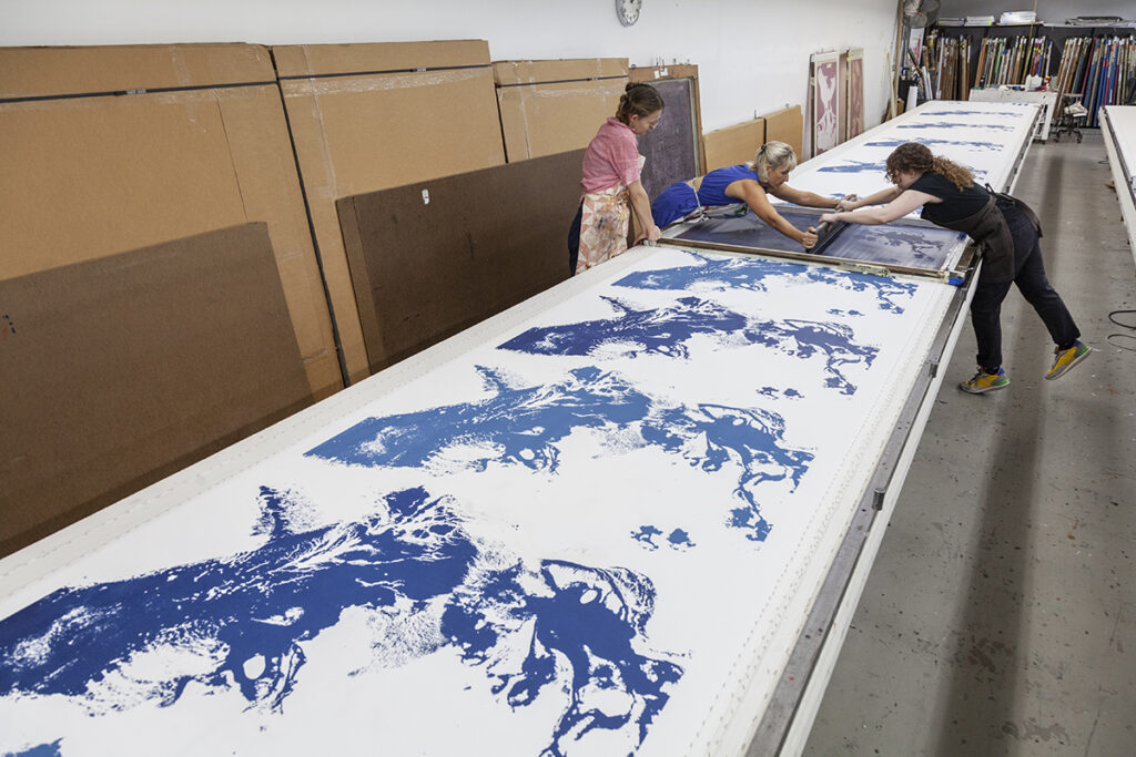 A photo of two women artists screenprinting from either side of a large table. They are printing a repeated abstract splash-like pattern in blue while a coworker looks on.