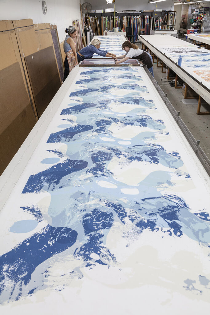 A photo of two women artists screenprinting from either side of a very long table. They are printing a repeated abstract splash-like pattern in blue while a coworker looks on.